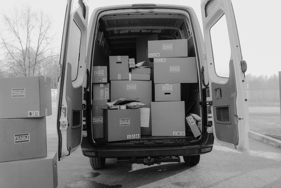 A small, vintage cream-colored van driving along a road during daytime, loaded with various household furniture and boxes. Inside the van’s open cargo area, there are wooden chairs, cardboard boxes,