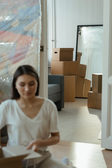 A man and a woman are inside a small flat, preparing for a home relocation. The woman, with long brown hair, is smiling and standing behind a partially opened cardboard moving box, which she appears t