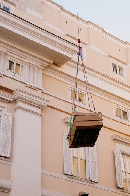 Close-up image of a yellow hoist trolley with a black hook hanging from the ceiling of an industrial warehouse. The hoist is suspended by two black cables and is positioned centrally against a backgro