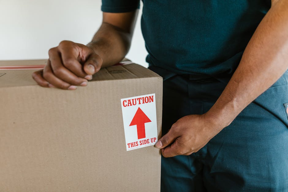 A close-up view of two large cardboard boxes packed for home relocation, placed on a wooden floor near a white wall. The top box, labeled with a white sheet of paper reading 'SUMMER CLOTHS,' contains 