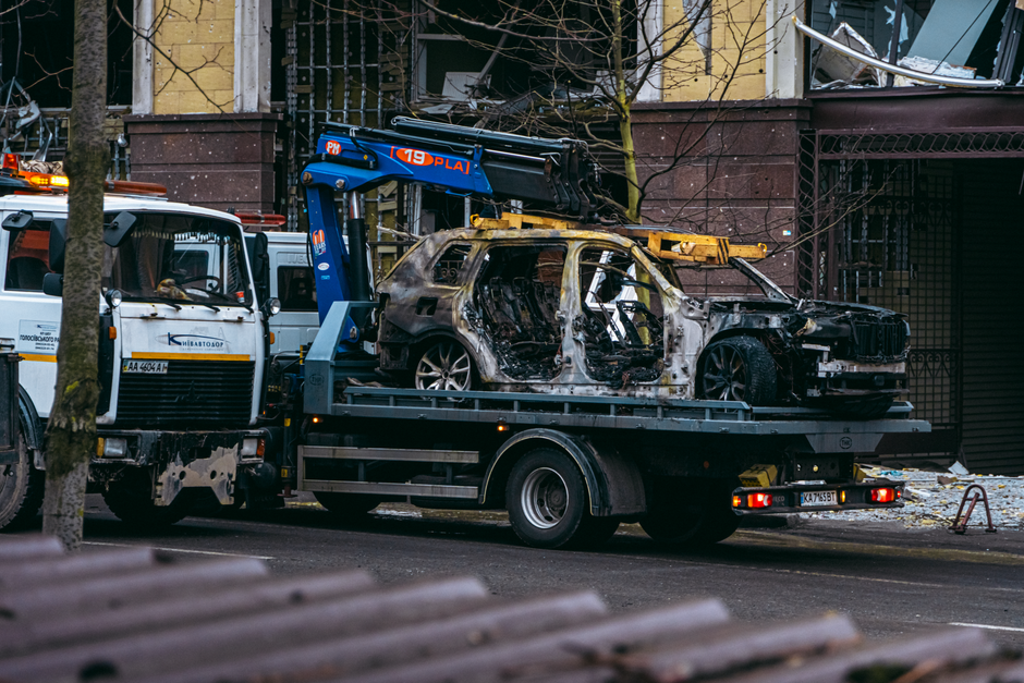 A damaged white sedan with extensive front-end impact sits atop a truck or trailer, positioned outside a dark wooden structure. The front of the car is crumpled and exposed, revealing internal compone