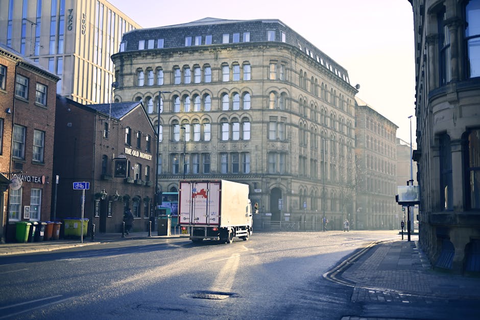 A white moving van with a closed cargo area is positioned on a city street, right outside a multi-storey, historic-style building with ornate architectural details and numerous arched windows. The van
